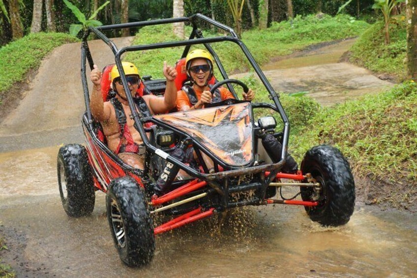 A red off-road vehicle with two people wearing helmets and life vests, driving on a muddy path surrounded by greenery. 4