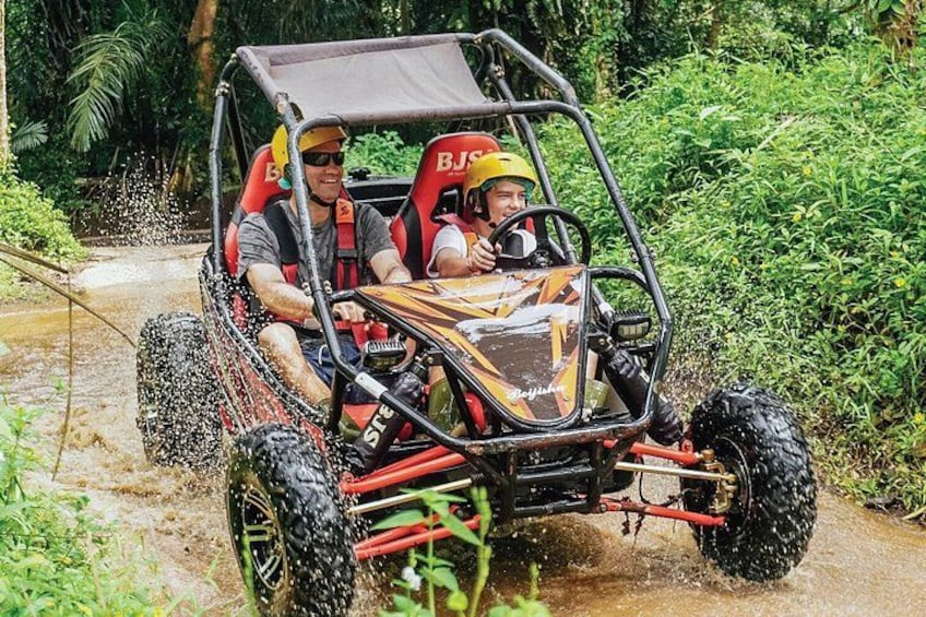 Un vehículo todoterreno de cuatro ruedas con dos pasajeros, uno de ellos con casco, circulando por un sendero embarrado rodeado de follaje. 5