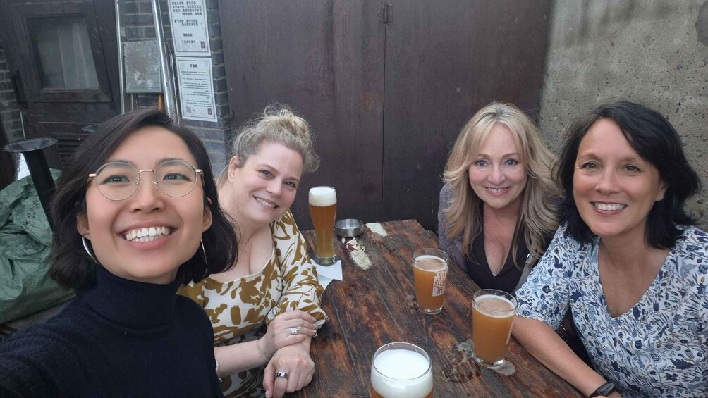 Four women sitting at a wooden table with drinks, in an outdoor setting with a brick wall and posters in the background. 5