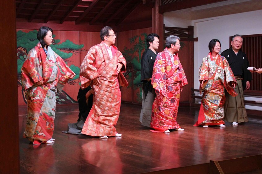 A group of people in traditional Japanese attire on a stage with a backdrop of painted scenery. 3