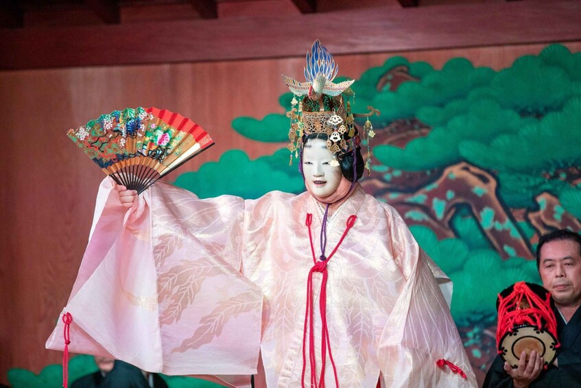 A person in traditional costume holding a fan and a drum, with a painted backdrop. 1