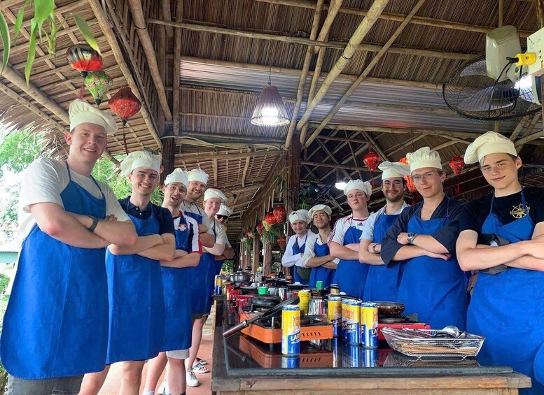 A group of chefs in blue aprons and chef hats standing in a covered outdoor kitchen area with cooking equipment and ingredients. 4