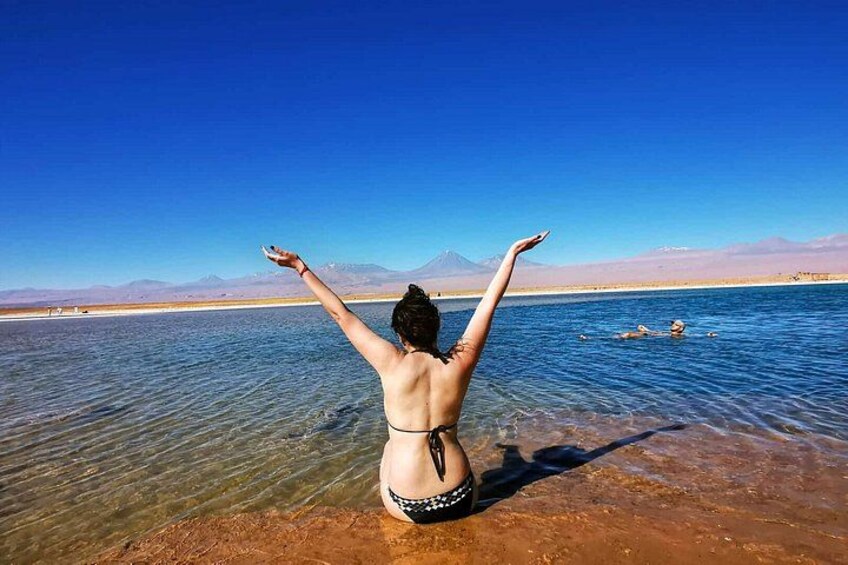 A person in a bikini standing in shallow water with arms raised, facing a clear blue sky and distant mountains. 1