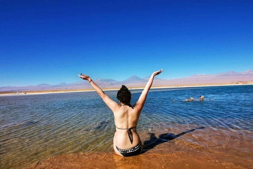 A person in a bikini standing in shallow water with arms raised, facing a clear blue sky and distant mountains. 1