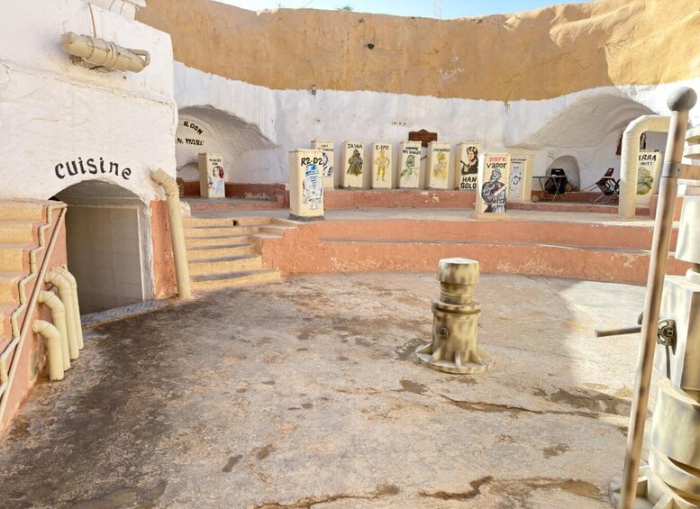 An ancient site with a central stone structure, steps leading to an entrance labeled 'cuisine,' and various artifacts displayed around the area. 5
