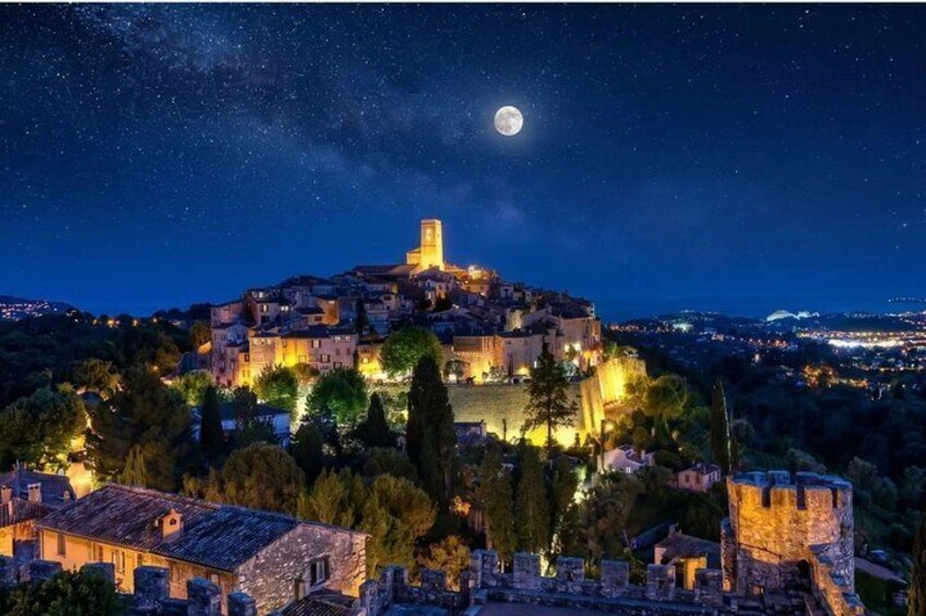 Une ville historique, dominée par une tour imposante, entourée d’arbres et bordée de bâtiments illuminés, sous un ciel étoilé où l’on peut apercevoir la lune. 4