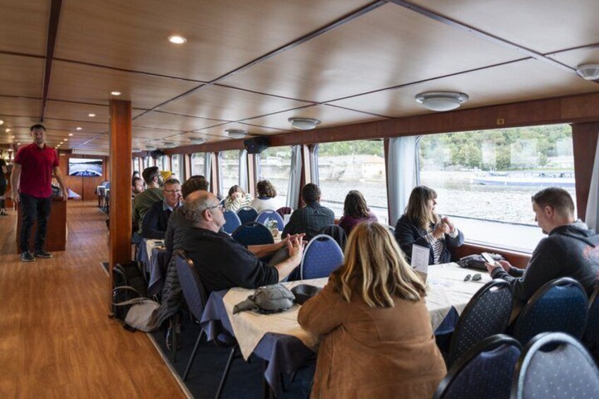 A dining area on a boat with people seated at tables, a waiter standing, and a view of the water outside. 4