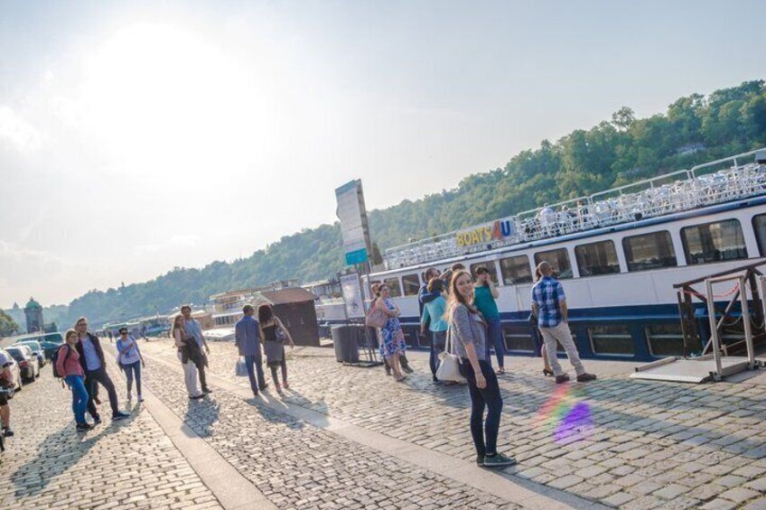 A group of people walking on a cobblestone path by a river, with a boat docked on the right. 3