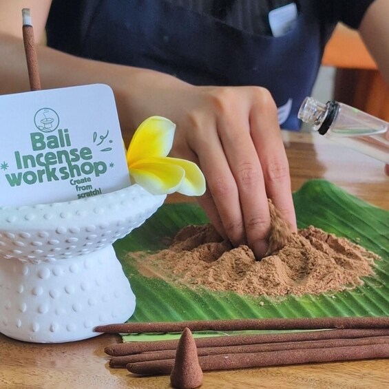 A person performing a Bali incense workshop, using a white bowl with a logo, a yellow flower, and brown incense cones on a green leaf. 1