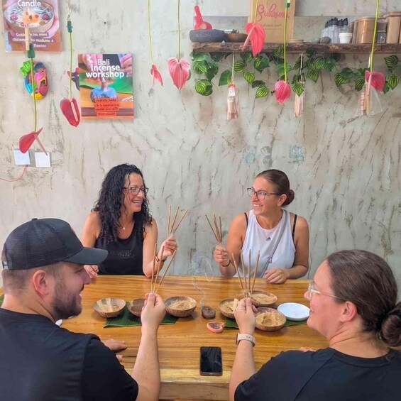 A group of people sitting around a table with incense sticks, a candle, and a book titled 'Bali Incense Workshop' on the wall. 5