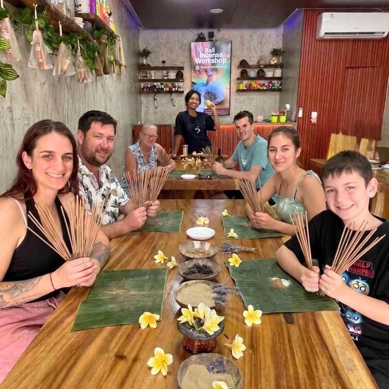 A group of people seated around a wooden table with incense sticks, flowers, and bowls, in a room with a poster and shelves in the background. 4
