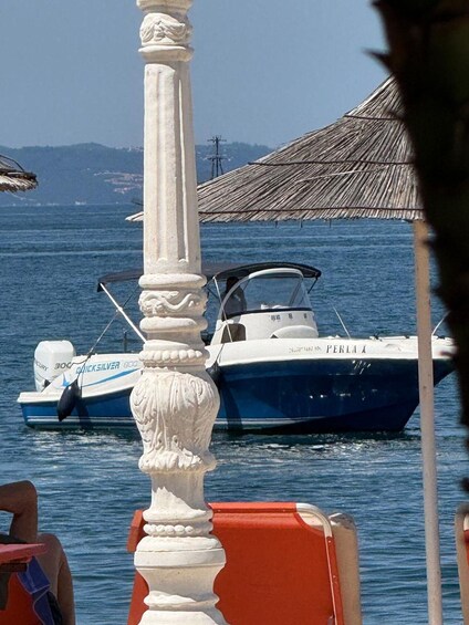 A boat named 'PERLA X' docked near a beach with a thatched umbrella and a white column in the foreground. 3