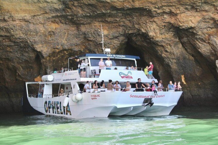 A boat named 'OPHELIA' with 'Sensational Sea Cruises' written on the side, docked in a rocky bay with a cave in the background. 1