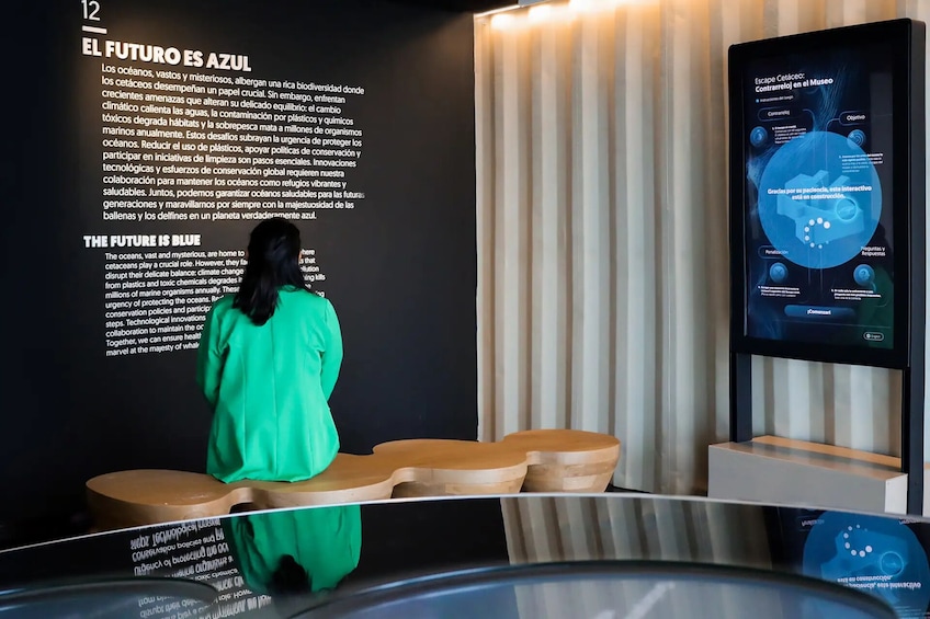 Una persona vestida de verde de pie frente a un expositor con información sobre la conservación marina y una pantalla digital con contenido interactivo. 2