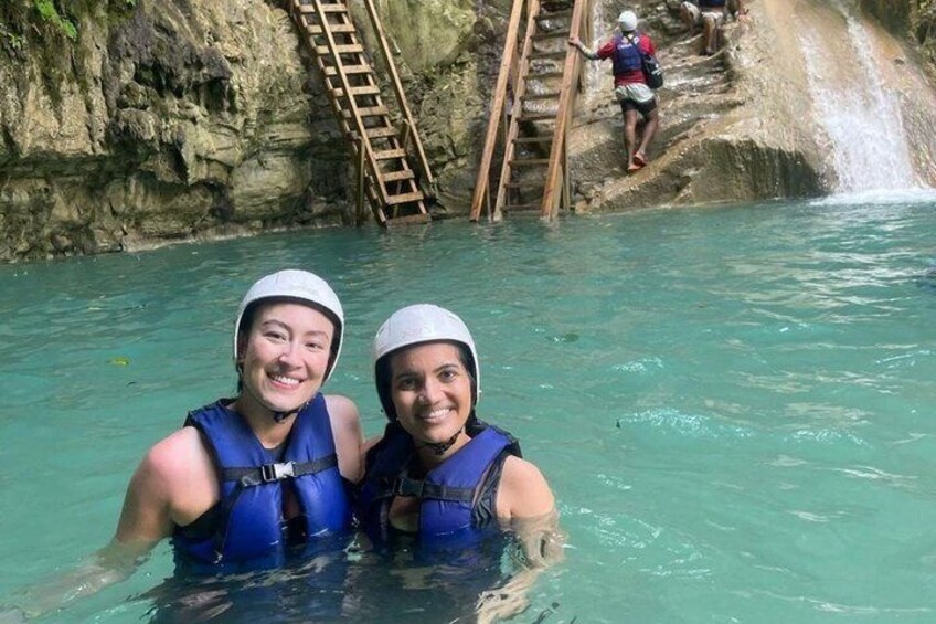 Two people in blue life vests in a clear water body with a rocky background and a wooden ladder. 1