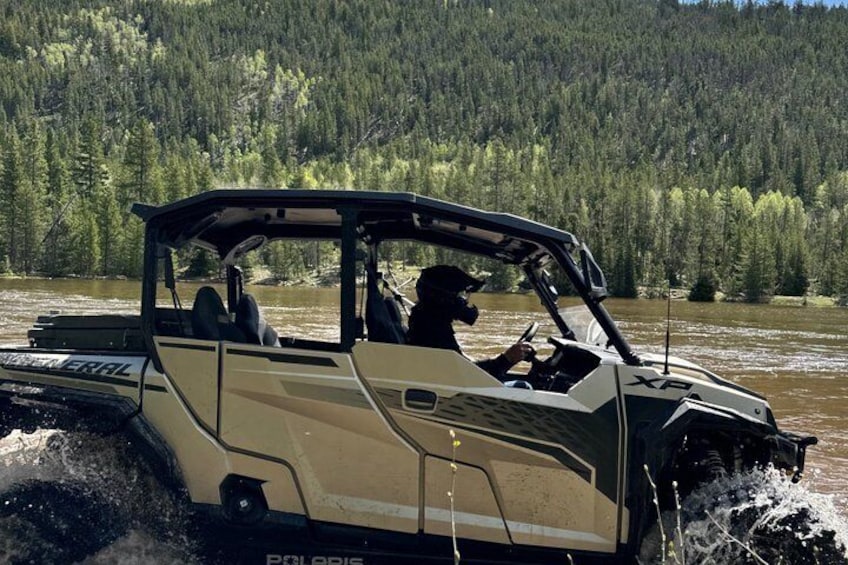 A beige off-road vehicle driving through a river surrounded by forest. 2