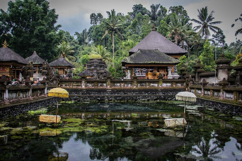 A traditional Balinese temple complex with thatched-roof structures, a central water body, and lush tropical vegetation. 2