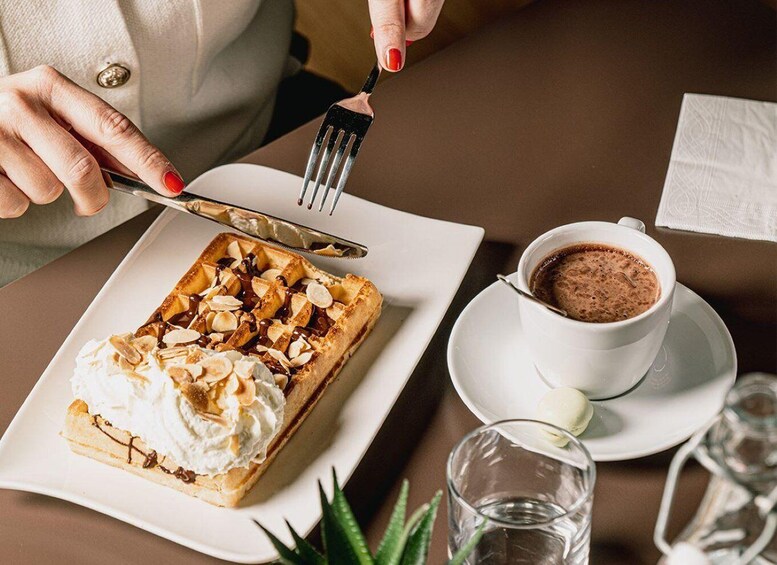 A person cutting a dessert with whipped cream and almonds, next to a cup of hot chocolate. 4