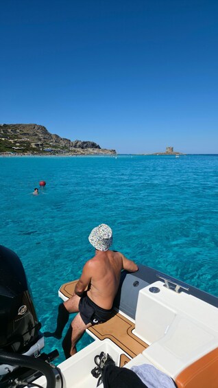 A person on a boat in clear blue waters with a rocky coastline and a tower in the distance. 1