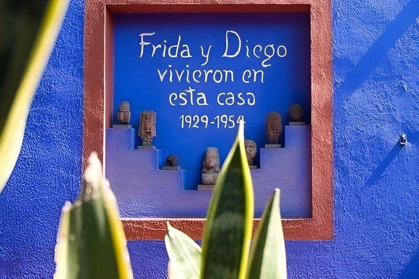 A blue wall with a plaque stating 'Frida y Diego vivieron en esta casa 1929-1954' and several sculptures. 1