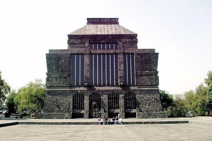 A large, rectangular building with a flat roof and vertical columns, surrounded by a paved area with trees. 3