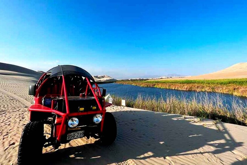 A red off-road vehicle parked on a sandy beach with a body of water and dunes in the background. 2