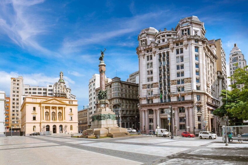 A city square with a prominent statue, classical architecture, and a clear blue sky. 1