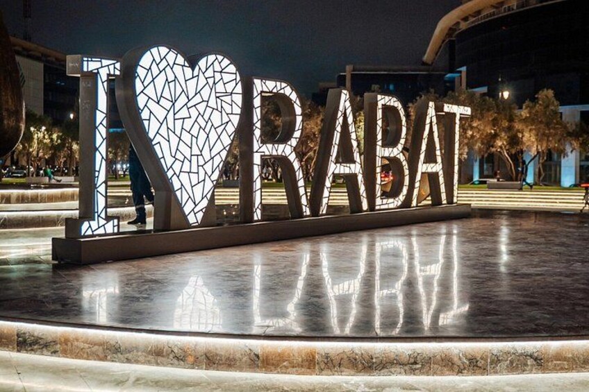 Een nachtelijke scène met verlichte letters die 'I LOVE RABAT' vormen op een reflecterend oppervlak, omgeven door een plein met bomen en een gebouw op de achtergrond. 5