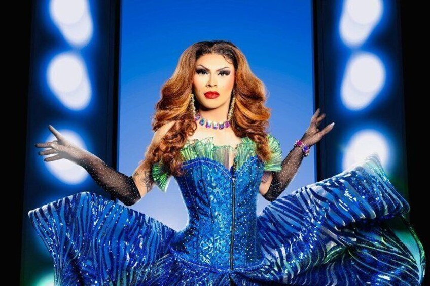 Una persona con un vestido azul con lentejuelas y falda amplia, de pie frente a un fondo azul con luces circulares. 1