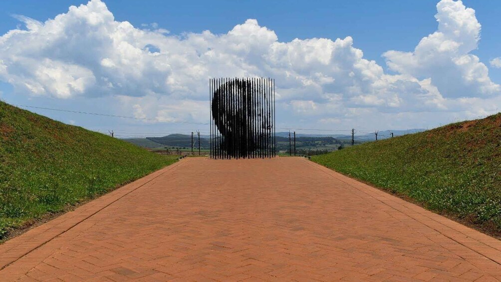 A paved pathway leading to a large, abstract sculpture in a rural landscape with hills and a clear sky. 1