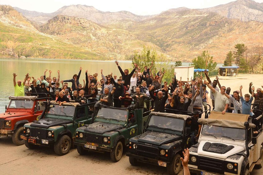 A group of people celebrating with raised hands, surrounded by off-road vehicles in a mountainous area with a lake. 5