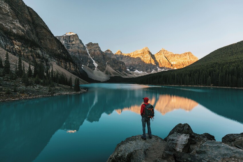 A person standing on a rock by a calm lake with mountains in the background. 2