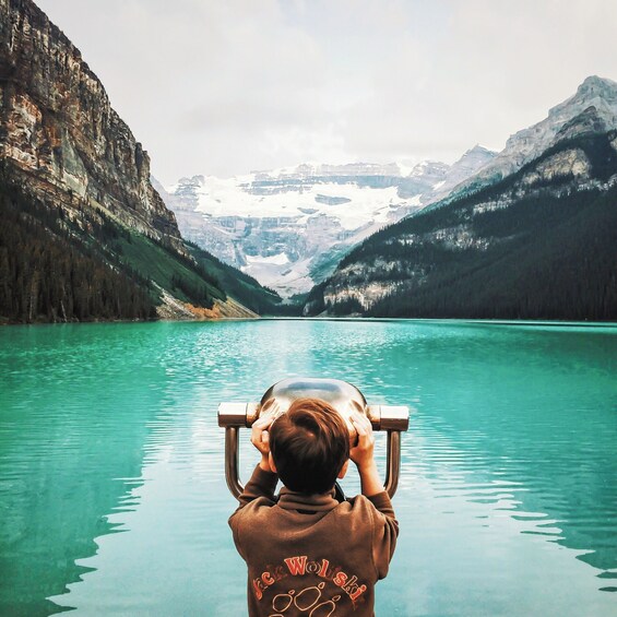 A person in a brown jacket with 'Jack Wolski' on the back, looking out over a turquoise lake with mountains in the background. 5