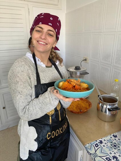 Eine Person steht in einer Küche, hält eine Schüssel mit Essen in der Hand und trägt einen Kochschürze. Auf der Arbeitsfläche liegen Kochutensilien und ein Topf. 3