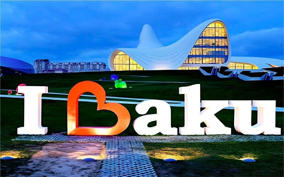 A modern building with a unique curved roof, surrounded by a well-maintained grassy area and a large illuminated sign that reads 'I Baku.' 5
