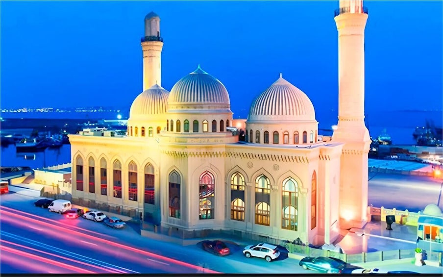 A mosque with two minarets and three domes, surrounded by a parking lot and a busy road. 3
