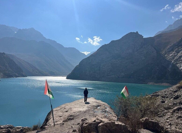 A person standing on a rocky outcrop overlooking a lake with mountains in the background. 1