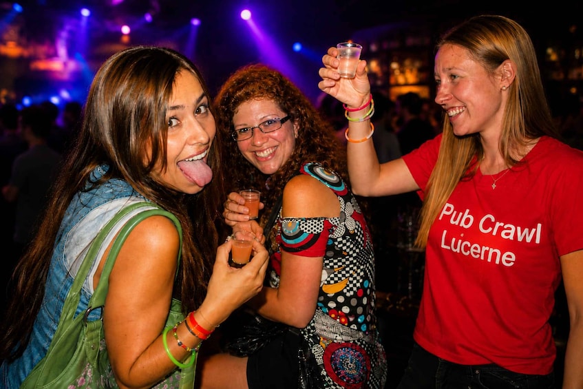 Three people at a social event, one wearing a red shirt with 'Pub Crawl Lucerne' text. 1