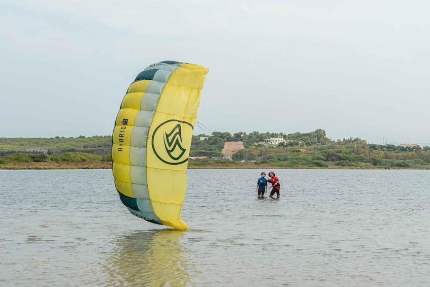 A parasail with a yellow and green sail, two people standing on a body of water, and a distant shoreline with buildings and greenery. 5