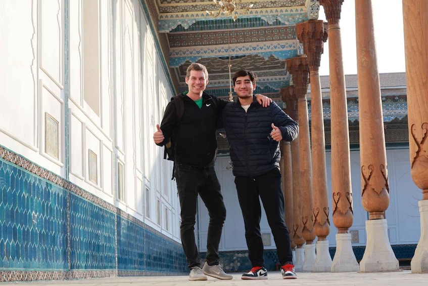 Two people standing in a corridor with ornate columns and patterned walls. 1