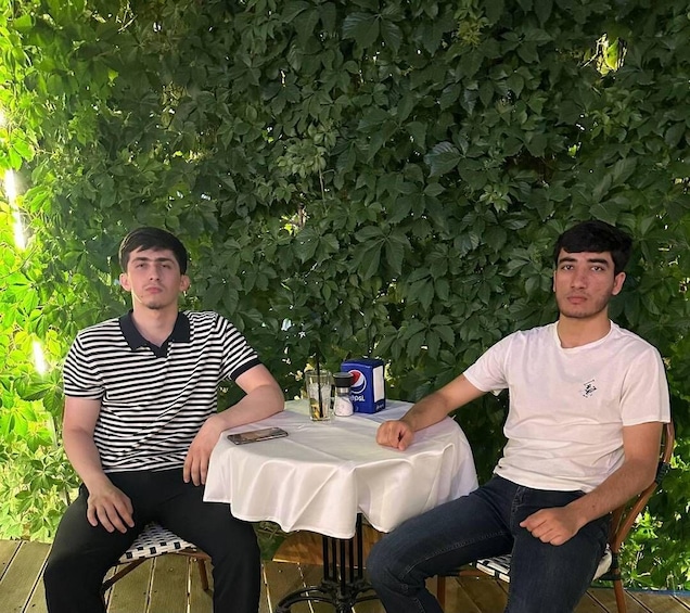 Two individuals seated at a table with a Pepsi can, surrounded by greenery. 3
