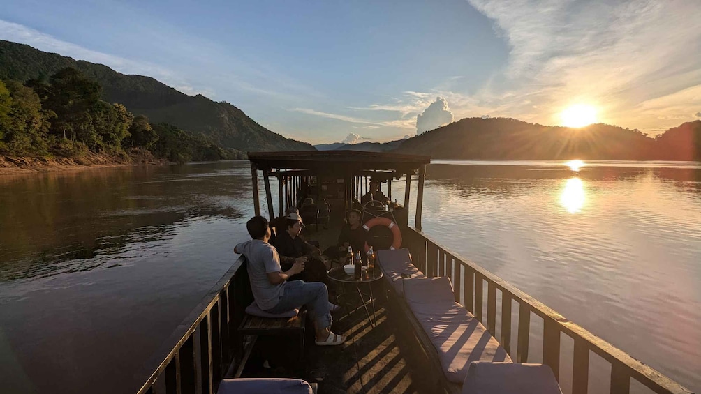 A boat with people sitting on the deck, a sunset in the background, and a calm river. 3