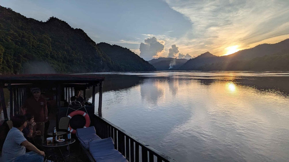 A boat with a covered deck, people sitting, and a calm river at sunset. 1