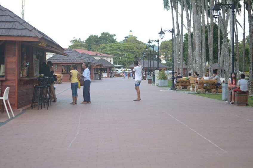 A paved walkway with wooden structures on the left, palm trees on the right, and people walking and sitting. 4