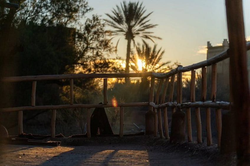Een houten hek met een poort, een palmpende en een ondergaande zon op de achtergrond. 2