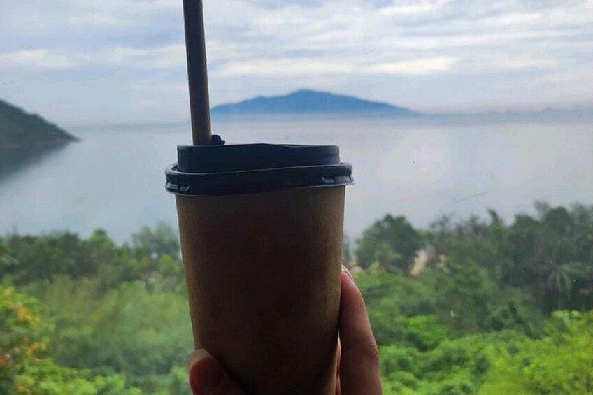 A hand holding a disposable coffee cup with a straw, overlooking a body of water and greenery. 2