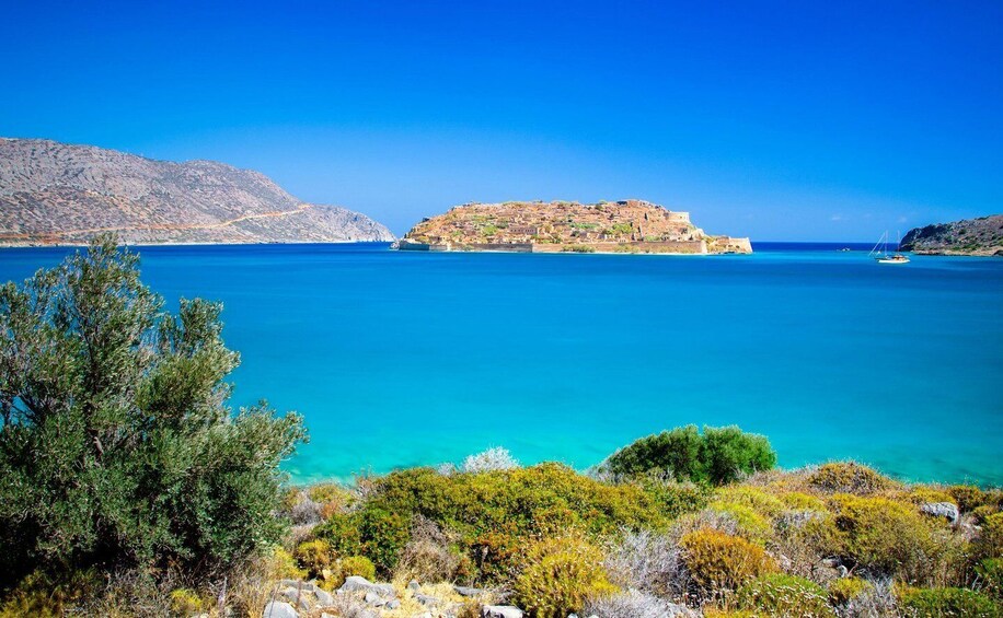 A coastal landscape with clear blue waters, rocky terrain, and a prominent hill with ruins. 1