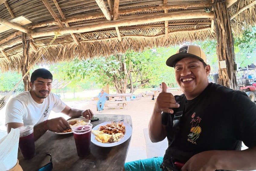 Two people sitting at a table with food and drinks, under a thatched roof structure. 2