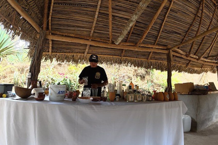 A person standing behind a table with various items, including bottles and containers, under a thatched roof structure. 4