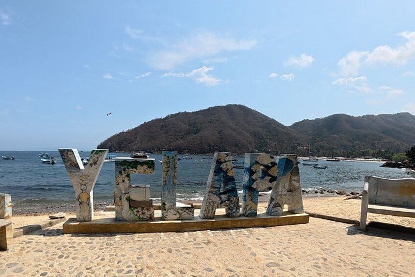 A beach with a large, abstract sculpture made of stone and metal, a sandy shore, and a mountainous backdrop. 1